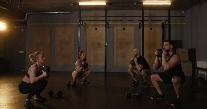 Four athletic people doing kettlebell squats and goblet squats during exercise in cozy modern fitness room. Group training warm-up the guys and girls in the gym