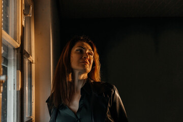 Pensive young caucasian female in leather jacket by window in moody lighting