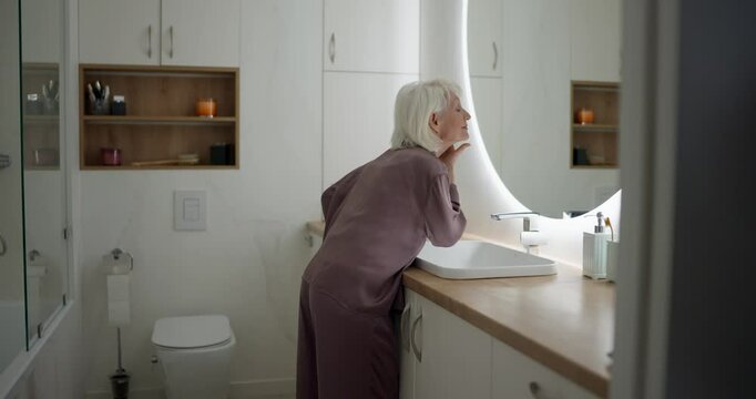 Woman senior in purple pajamas examining her face and doing facial massage in front of mirror in the morning in modern bathroom. Morning of an elderly woman