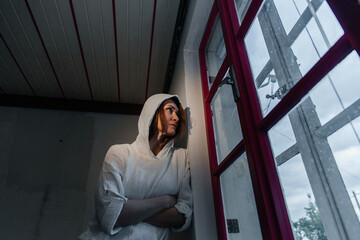 Caucasian female in hoodie gazing out of red-edged window in dimly lit room