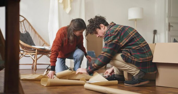 Focused guy and his girlfriend cutting wrapping paper with scissors and wrapping dishes in it while moving house