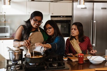 Three young South Asian women cook a traditional meal indoors in a modern kitchen. They wear festive clothing and stir ingredients in a pan on the stove during the daytime.