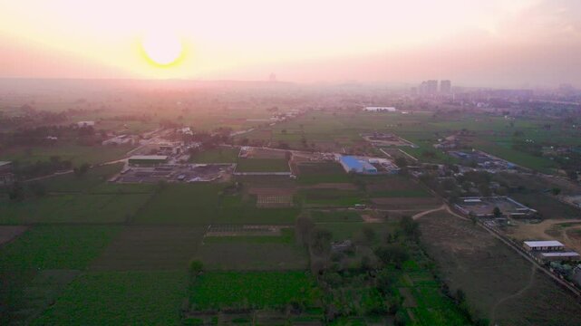 Aerial drone shot flying forward over green fields farms with houses showing outskirts, rural and industrial areas near Gurugram Haryana Delhi with skyscrapers in distance