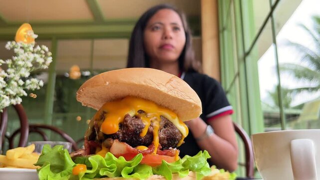 young woman drinking milkshake against background of huge juicy burger and sauces on table in restaurant