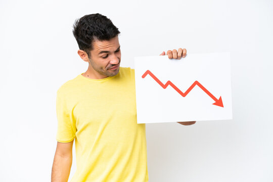 Young handsome man over isolated white background holding a sign with a decreasing statistics arrow symbol with sad expression