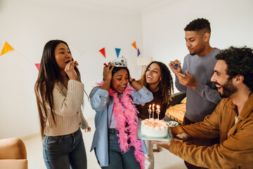 A multiracial group of friends celebrates a birthday indoors. One woman wears a crown and pink feather boa and looks surprised as a cake with lit candles is brought to her while others cheer.