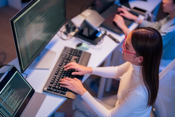 asian female programmer sits a dark tech workspace at night, fully focused on developing an online...