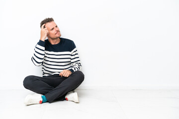 Young man sitting on the floor isolated on white background having doubts and with confuse face expression