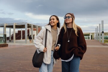 On a city square with a wooden floor, two multinational women in their 20s stroll, smile, and chat on a cloudy day. Concept of a weekend walk, a meeting with a close friend.