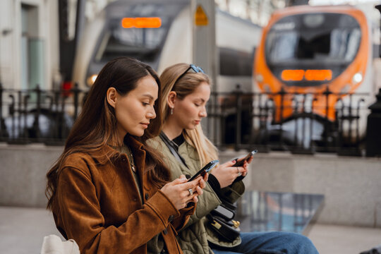 Two young White women sit at a train station outdoors, using smartphones while waiting for the train. They wear casual clothes.