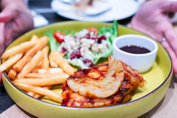 Delicious grilled chicken steak topped with pineapple, served with crispy fries, fresh salad, and a side of BBQ sauce on a yellow plate.