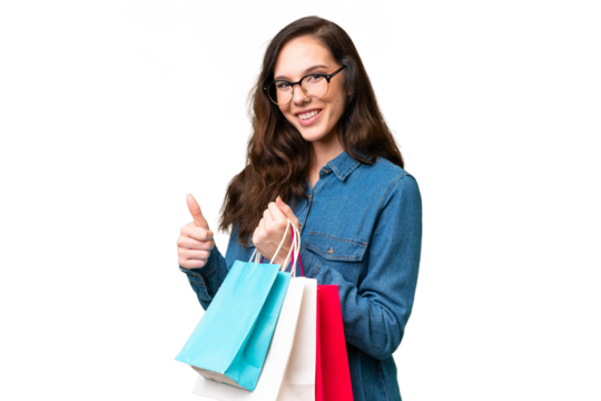 Young caucasian woman over isolated background holding shopping bags and with thumb up - Powered by Adobe