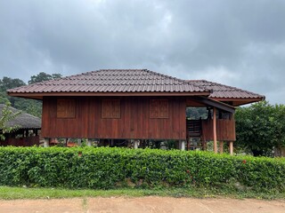 A traditional wooden Thai house with a brown tiled roof, elevated on stilts, and surrounded by lush green hedges and trees, under an overcast sky.