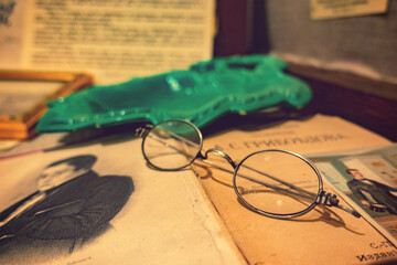 Vintage still life with antique round wire-rimmed glasses resting on yellowed book pages, green glass ashtray, and old newspaper with portrait.