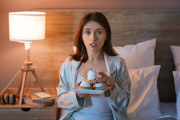 Young shocked pretty woman with cakes on bed in bedroom at night
