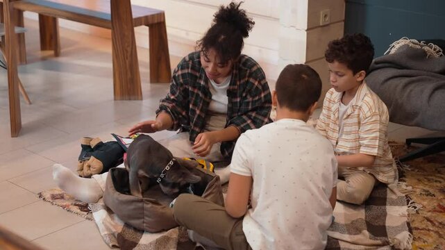 A light brown-skinned family, including a mother and her two school-aged sons, sits on the floor and pets a dog. A concept of people relationships, leisure time, and bonding,