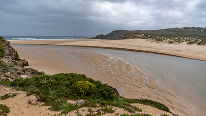 Praia da Amoreira im Fr&uuml;hling bei Ebbe, langgezogener Fluss flie&szlig;t bei Ebbe durch kleine Sandd&uuml;nen, umrahmt von gr&uuml;nem Bewuchs, dunkle Wolken am Himmel, 