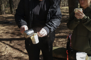Outdoor morning ritual in a pine forest as a couple shares a quiet drink; one pours fresh coffee into a white enamel mug while the other clutches their own cup, enjoying the warmth