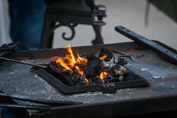 Close-up of a traditional blacksmith forge with burning coals, tools on the metal surface, and people standing nearby during an outdoor event.
