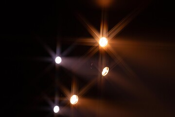 Multiple bright stage lights form vivid starburst patterns against a dark background, creating an...