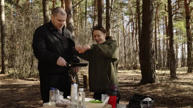 woman breaking eggs into a hot pan while a man holds the skillet steady over a makeshift camping table
