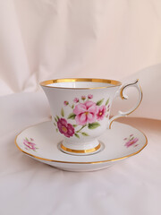 Fototapeta premium A porcelain teacup with floral design and gold trim sits on a matching saucer against a soft, light-colored fabric background