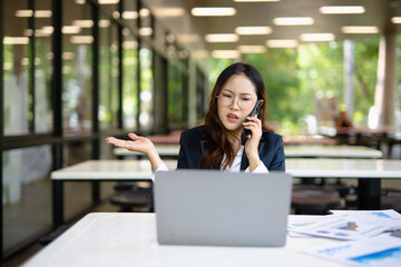 Fototapeta premium Asian businesswoman working on financial market research using laptop and digital tablet, stressed about digital investment in office.