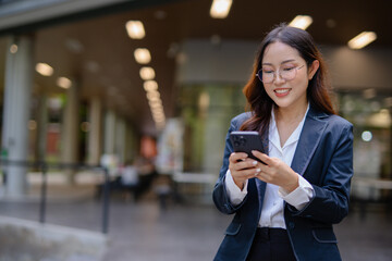 Asian businesswoman in suit using mobile phone while standing in front of modern business building.