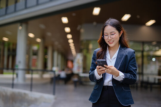 Asian businesswoman in suit using mobile phone while standing in front of modern business building.