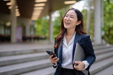 Asian businesswoman in suit using mobile phone while standing in front of modern business building.