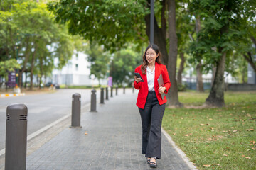 Smiling Asian businesswoman in suit holding coffee cup and documents walking in front of modern business building