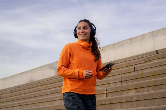 Young brunette woman in her early 20s, fit and sporty, wearing leggings and a top, using her phone with headphones on, selecting music to get motivated before her workout.