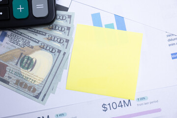 Hundred-dollar bills, a calculator, and a yellow sticky note placed on financial documents showing charts and figures.
