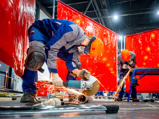 Two workers in protective gear cutting metal pipes with angle grinders, sparks flying, in a safety-zoned industrial workspace. Metalworking. Grinding. Fabrication. Construction.