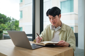 Focused professional man taking notes with headphones around neck near large window.