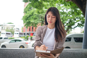 Fototapeta premium Thoughtful Asian woman writing in notebook while sitting outdoors in urban setting.