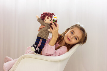  Beautiful little girl in pink dress sits on white chair with soft knitted toy in hands, selective focus