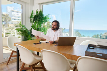 Bearded Man Working Remotely with Laptop in Modern Home Office Overlooking Scenic Ocean View