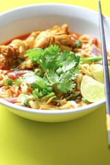 A bowl of spicy noodle soup with chicken, garnished with fresh cilantro, lime wedges, and chopped vegetables, served with black chopsticks. The vibrant colors and presentation are visually appealing.