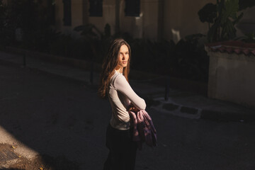 A young woman in a white top stands in warm evening sunlight, holding a pink blazer. Her expression...