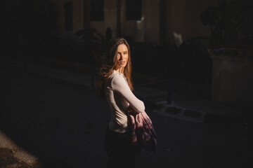 A young woman in a white top stands in warm evening sunlight, holding a pink blazer. Her expression...