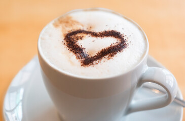 White cup on saucer with a chocolate powder heart on the coffee milk foam. On light wooden background. Viewed from above at an angle, focus on the powder