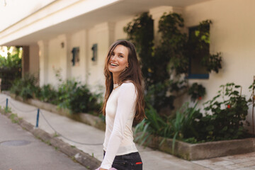 Fototapeta premium Happy woman walking outdoor on the street turn around, smiling. Brunette long hair girl wear long sleeve top and hold stylish pink blazer. Gold necklace. Positive emotions, fashion outfit.