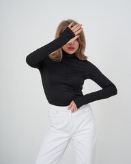 Woman in black turtleneck and white pants covers face with one hand, standing in a stylish pose against a plain backdrop. Her hair is loose and softly styled
