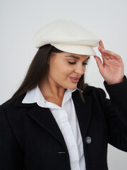 Smiling woman wearing a white beret and black coat, adjusting her hat. The woman has long dark hair and wears a white shirt underneath the coat, enjoying a stylish and classic look