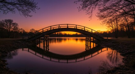 Fototapeta premium Serene sunset over a picturesque wooden arched bridge mirrored in still water, framed by leafless trees