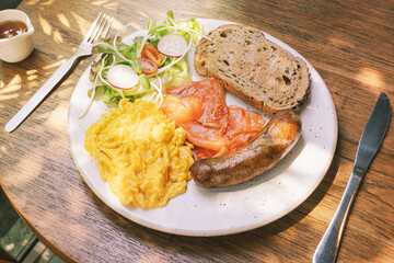 Delicious breakfast plate featuring scrambled eggs, smoked salmon, sausage, fresh salad, and whole grain toast served in a cozy outdoor cafe setting. Rustic English breakfast on a wooden table in