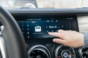 A closeup view of a cars modern dashboard showcasing a sleek digital display alongside well placed radio controls. The passenger controls the radio on the car's touchscreen with his finger.