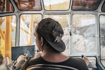 Crane operator at work in the cabin, close-up view from the rear. Crane operator focused on safely loading coal from a barge at a bustling dock situated along the river. wearing a cap, controls the