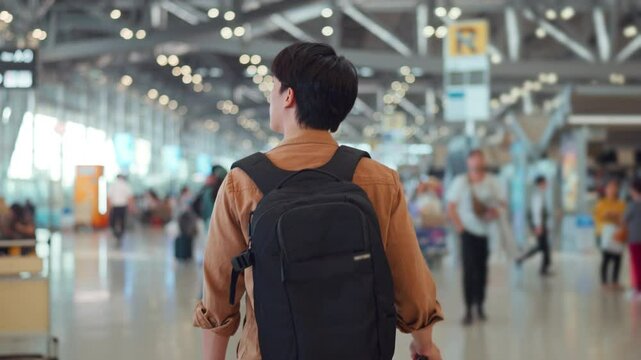 Rear view - Asian man tourist passenger with backpack walking to find his way to the boarding gate inside the airport terminal. Tourist journey concept